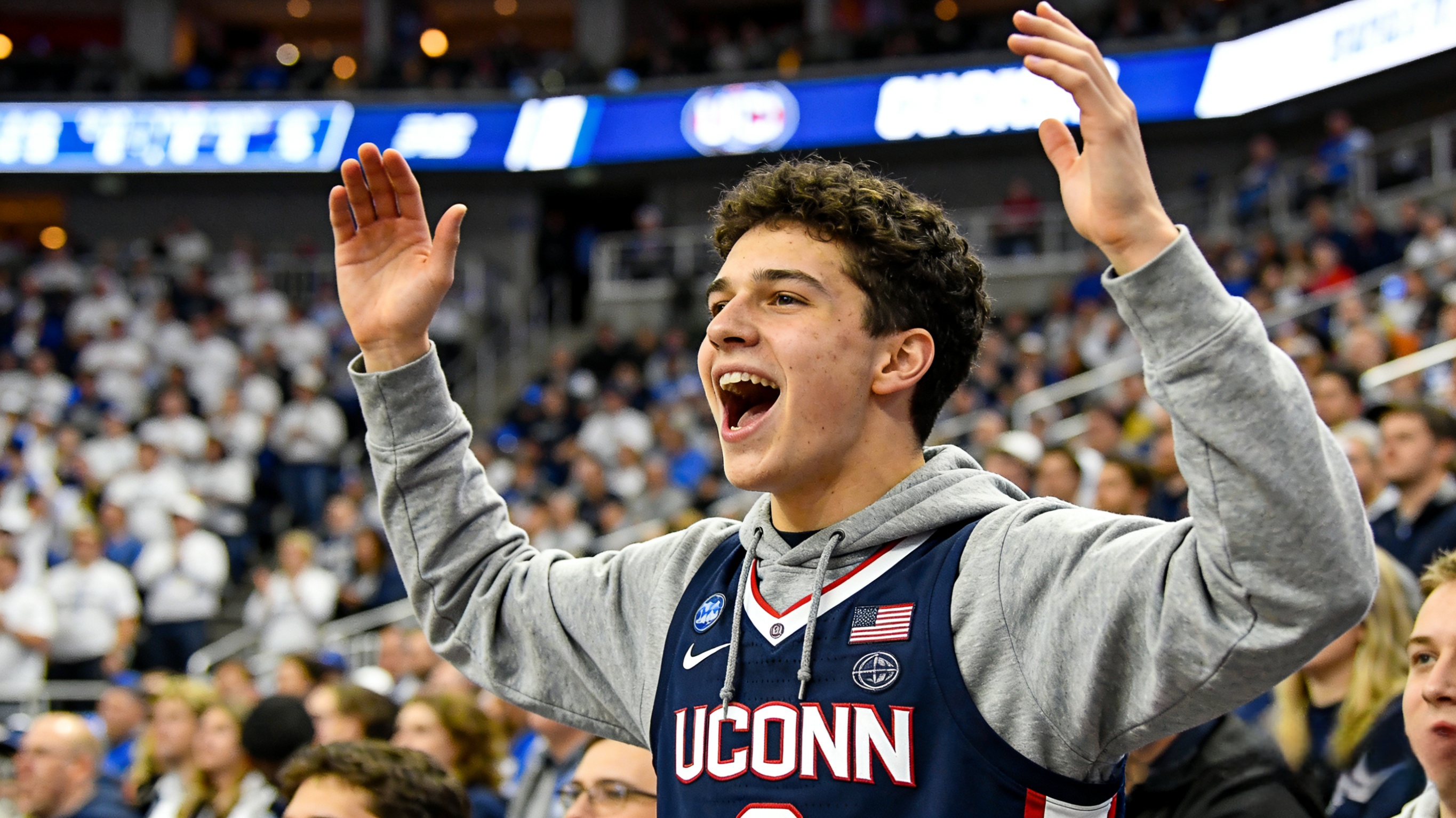 A fan wearing a UConn basketball jersey cheering at an indoor basketball arena.