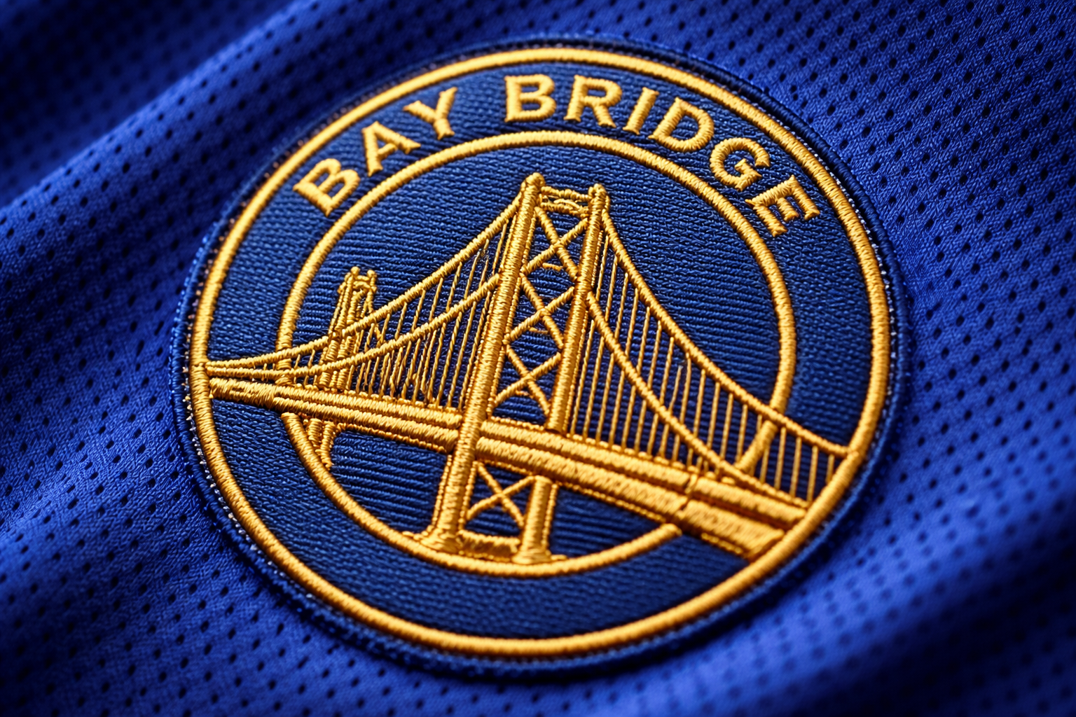 Warriors Jersey Guide: We Believe vs. Steph Curry Era 3 Macro close up of the gold stitched Bay Bridge logo on a blue Warriors jersey